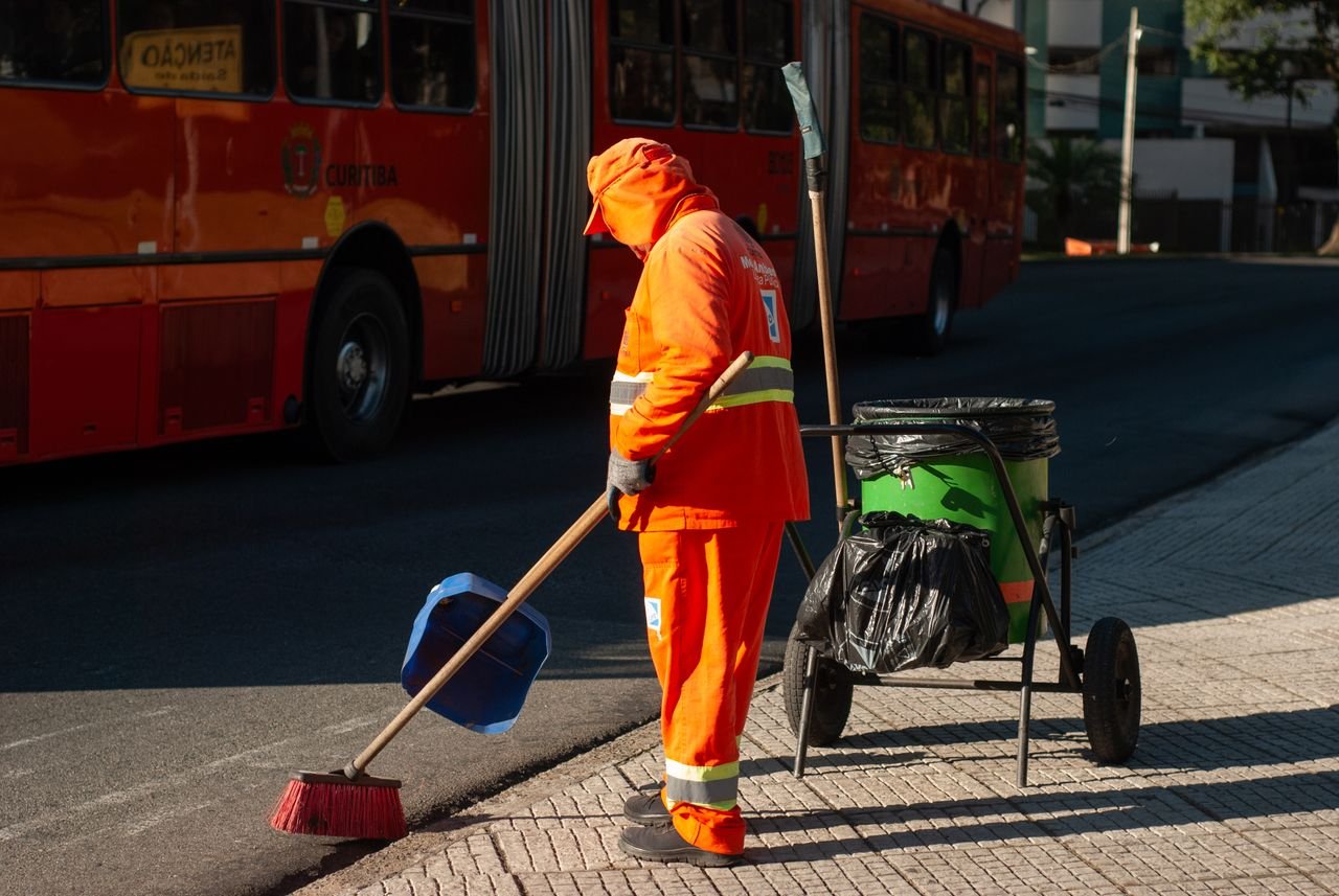 Street cleaner in bright orange uniform sweeping sidewalk near public bus in city setting.